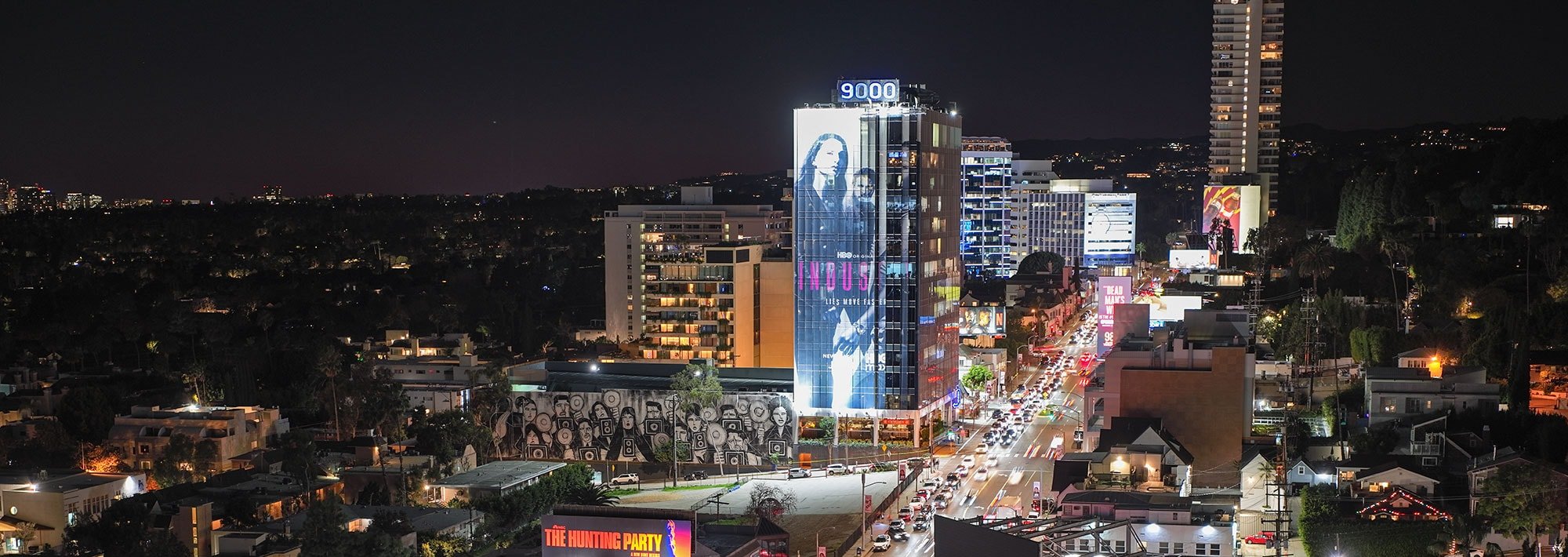An aerial nighttime view of the Sunset Strip in West Hollywood, the 9000 building lit against the dark hills, traffic streaming down Sunset Boulevard, billboards and city lights stretching toward the horizon