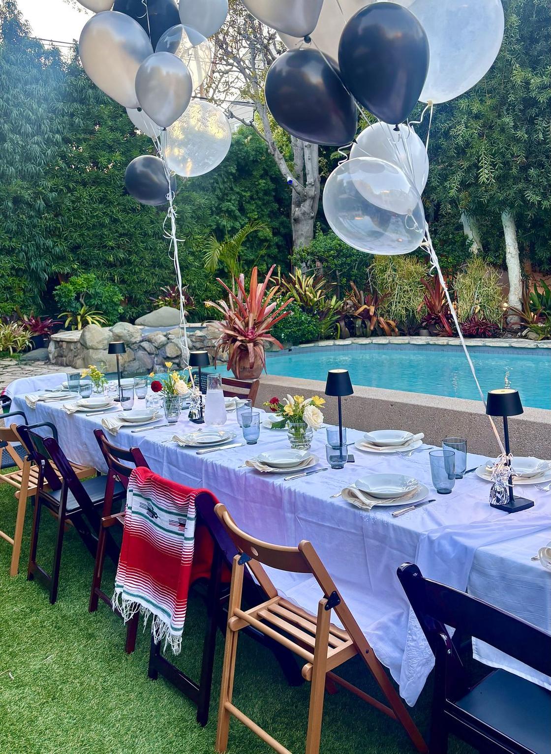A long table set for a dinner party in a Los Angeles backyard, plates and glasses laid, chairs empty, balloons overhead and the pool beyond